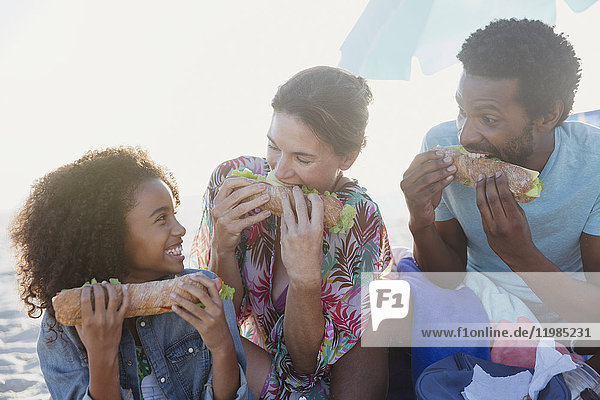 Verspielte multiethnische Familie mit Baguette-Sandwiches am Strand