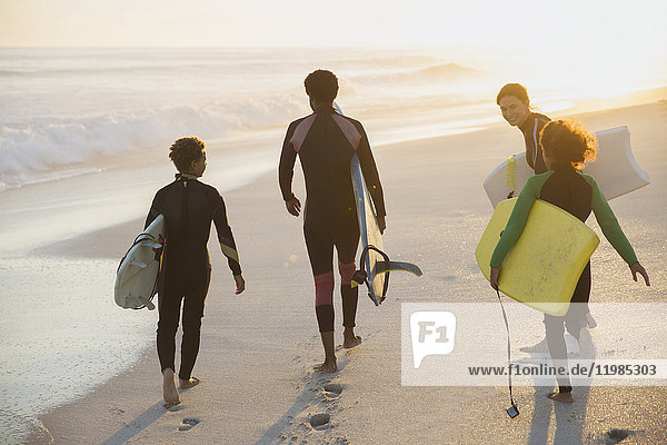 Multi-ethnic family carrying surfboard and boogie boards on sunny summer sunset beach