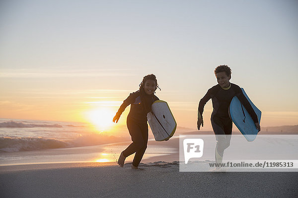 Brother and sister in wet suits running with boogie boards on summer sunset beach