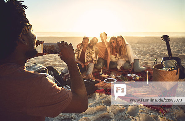 Young man with camera phone photographing friends enjoying picnic on sunny summer beach
