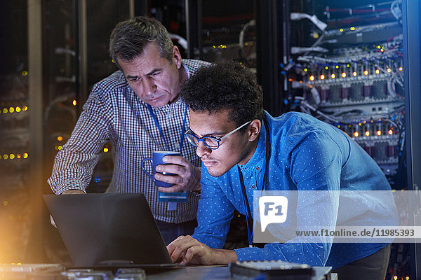Focused male IT technicians working at laptop in dark server room