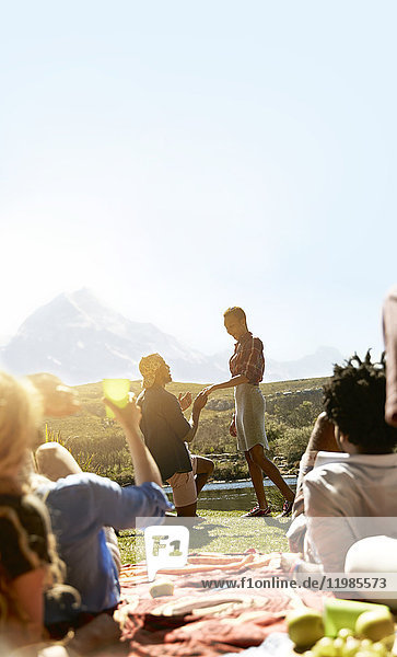 Freunde beobachten den jungen Mann  der der Frau bei einem sonnigen Sommerpicknick einen Antrag macht