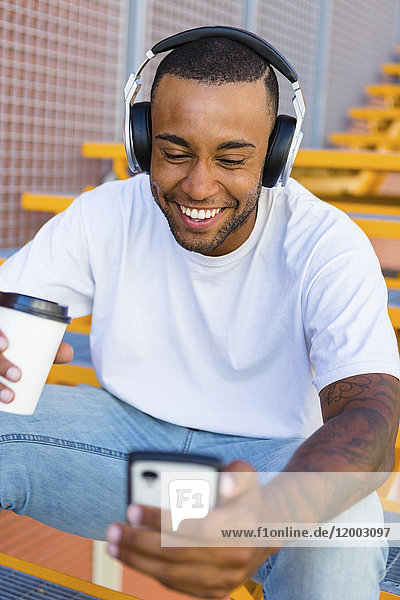 Laughing young man with headphones and coffee to go sitting on stairs looking at smartphone