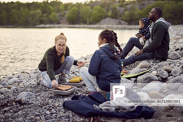 Mutter beim Zubereiten von Speisen auf dem Grill inmitten einer Familie  die während des Campings auf Steinen am Strand sitzt.