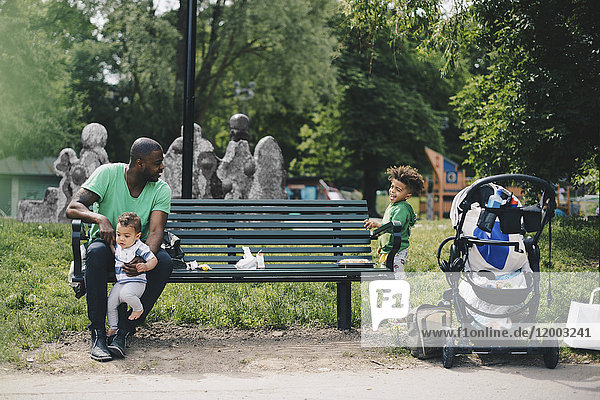 Father holding toddler and looking at son while sitting on bench at park