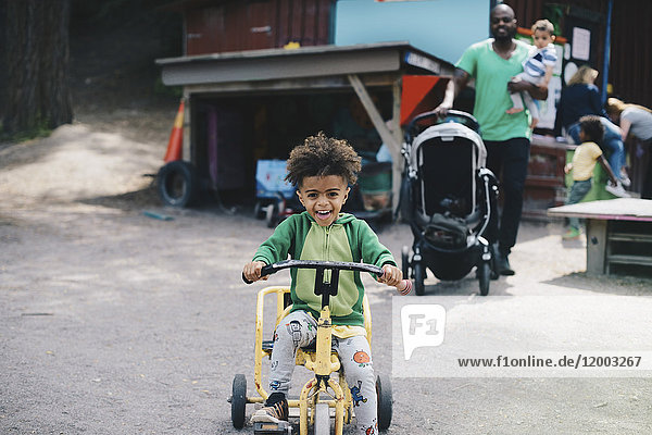 Fröhlicher Junge auf dem Dreirad,  während Vater und Bruder im Hintergrund laufen.