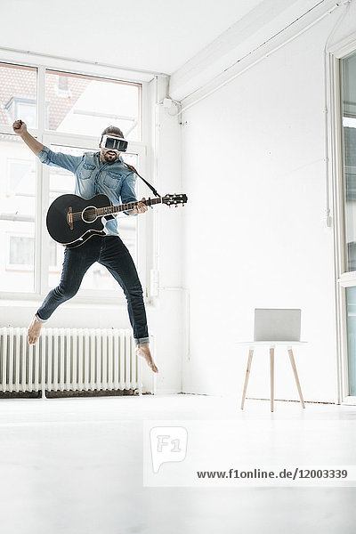 Mann mit Gitarre  der eine Virtual Reality Brille trägt  die in einem Loft in die Luft springt.