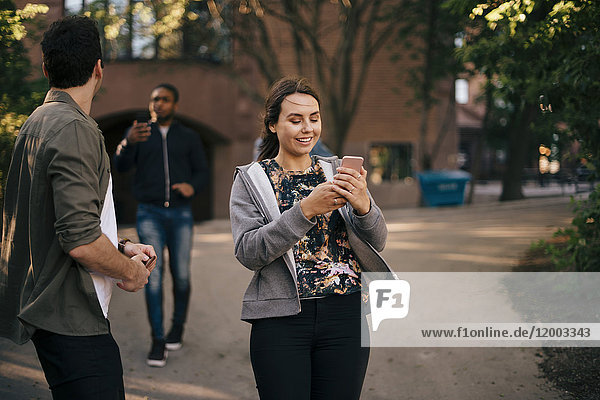 Lächelnde Frau mit Smartphone beim Spaziergang mit männlichen Freunden auf dem Fußweg