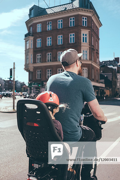 Rückansicht des Vaters beim Radfahren  während die Tochter auf dem Rücksitz in der Stadt sitzt.