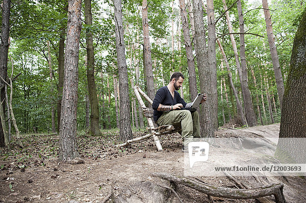 Mann sitzend auf selbstgemachtem Holzstuhl im Wald mit Laptop