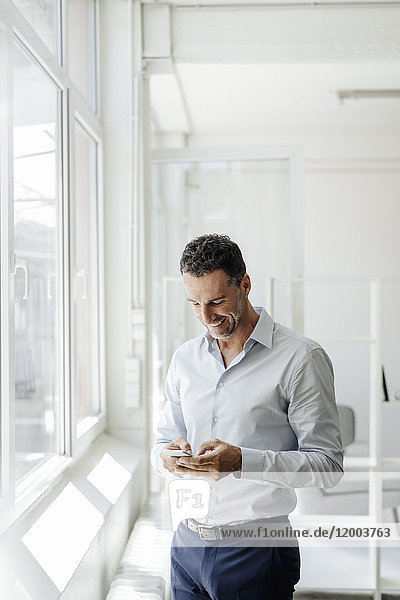 Smiling businessman in office at the window looking at cell phone