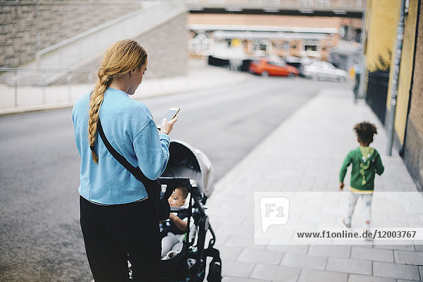 Rückansicht der Mutter mit Handy und schiebendem Kinderwagen beim Spaziergang mit dem Sohn auf dem Bürgersteig in der Stadt