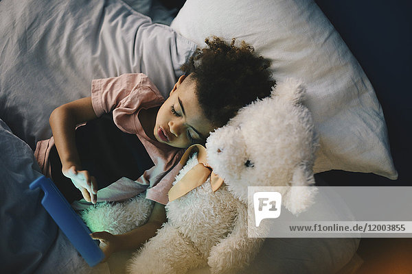 Hochwinkelansicht des Jungen,  der mit Teddybär liegt,  während er zu Hause mit einem digitalen Tablett auf dem Bett liegt.