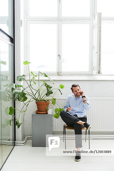 Volle Länge des Geschäftsmannes  der auf dem Handy spricht  während er bei einer Topfpflanze im Kreativbüro sitzt.