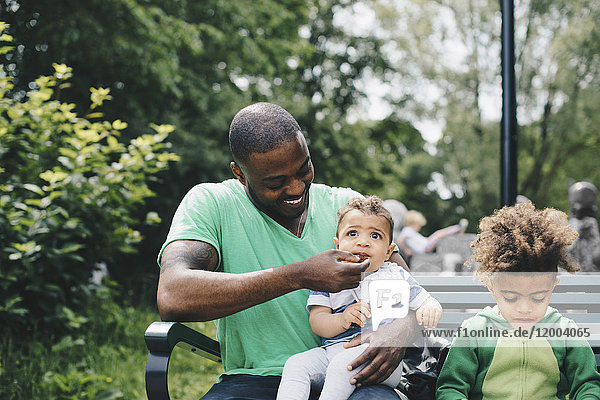 Lächelnder Vater füttert den Jungen  während er mit seinem Sohn auf der Bank im Park sitzt.