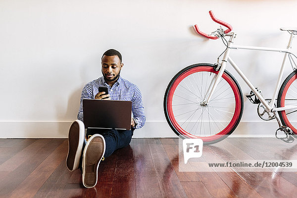 Mann auf Holzboden sitzend mit Laptop und Handy und Fahrrad neben ihm