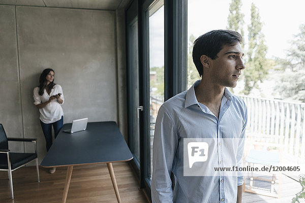 Young man looking out of window with woman in background