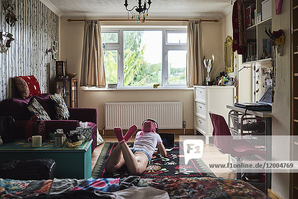 Junge Frau mit rosa Haaren  die auf einem Teppich liegt  trägt Kopfhörer und benutzt den Laptop zu Hause.