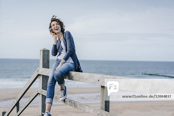 Glückliche Frau sitzt auf einem Geländer am Strand und hört Musik.