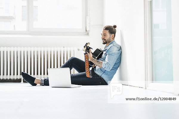 Mann sitzt mit Laptop auf dem Boden und spielt Gitarre