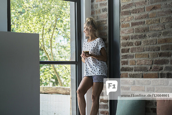 Reife Frau steht am Fenster und trinkt Kaffee.
