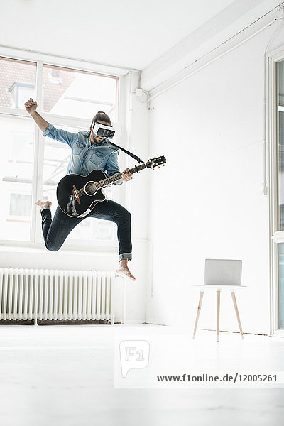 Mann mit Gitarre  der eine Virtual Reality Brille trägt  die in einem Loft in die Luft springt.
