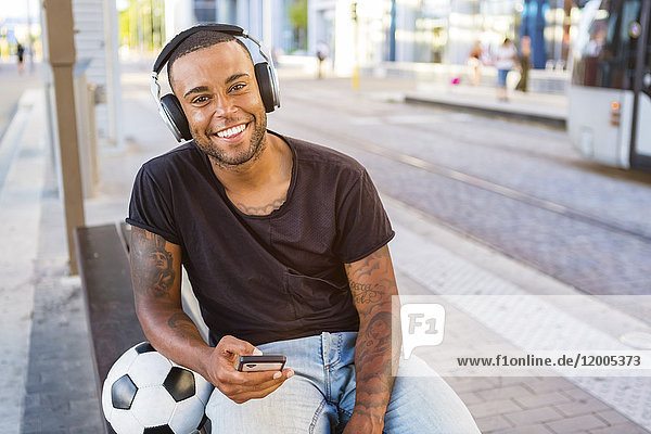 Porträt eines lächelnden jungen Mannes mit Fußball  Kopfhörer und Handy an der Straßenbahnhaltestelle