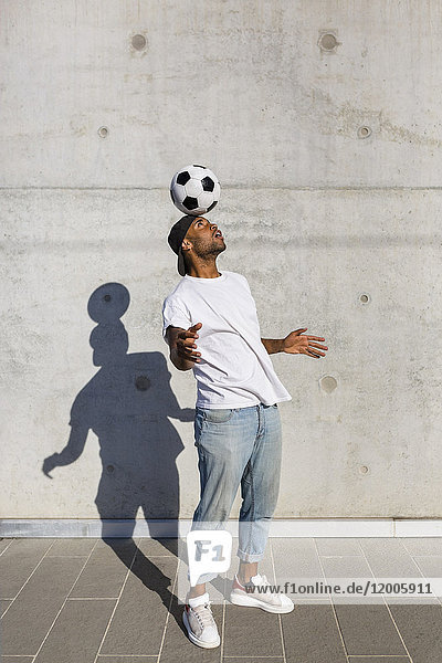 Young man balancing soccer ball on his head in front of concrete wall