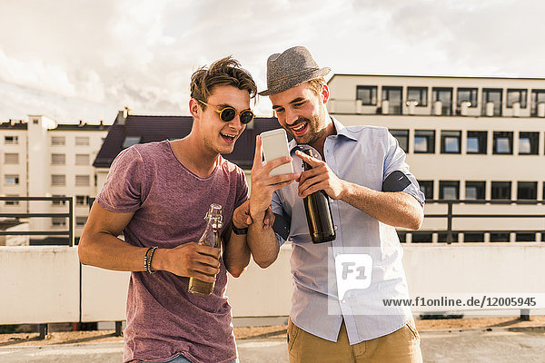 Two friends with beer bottles and cell phone on rooftop