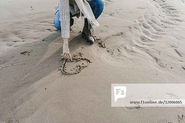 Frau kauert am Strand und zeichnet ein Herz im Sand.