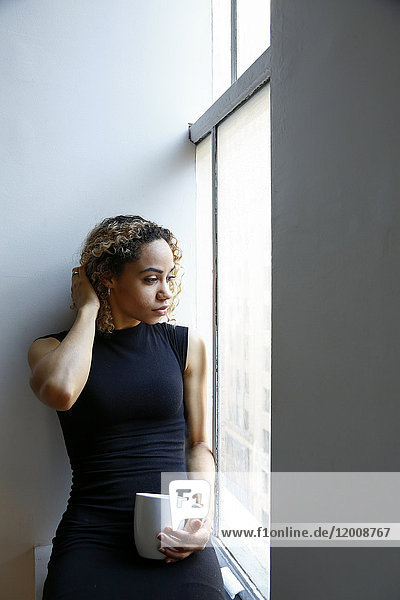 Pensive mixed race woman sitting on windowsill drinking coffee