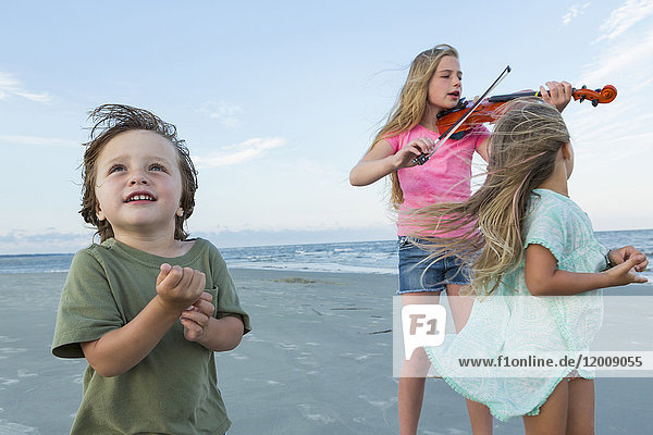 Kaukasisches Mädchen spielt Geige am windigen Strand neben Bruder und Schwester