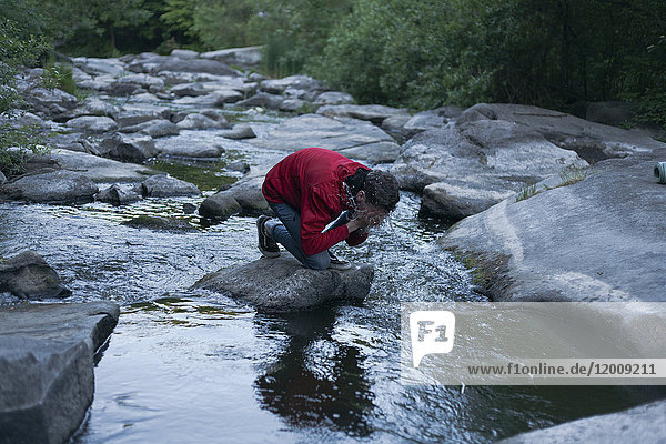 Kaukasischer Mann kniet auf einem Felsen im Fluss und spritzt sich Wasser ins Gesicht