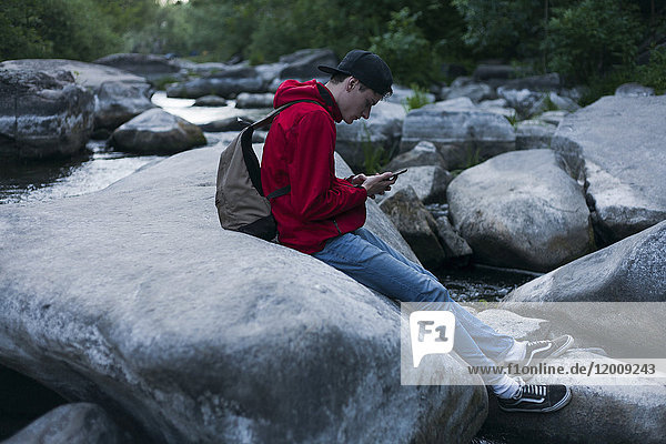 Kaukasischer Mann sitzt auf einem Felsen im Fluss und schreibt eine SMS mit seinem Handy