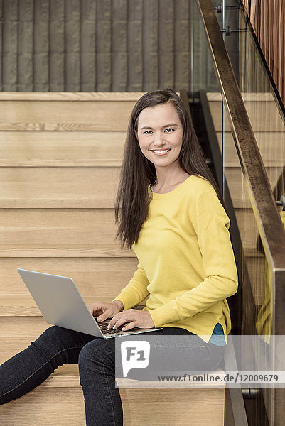 Lächelnde gemischtrassige Frau  die auf einer Treppe sitzt und einen Laptop benutzt