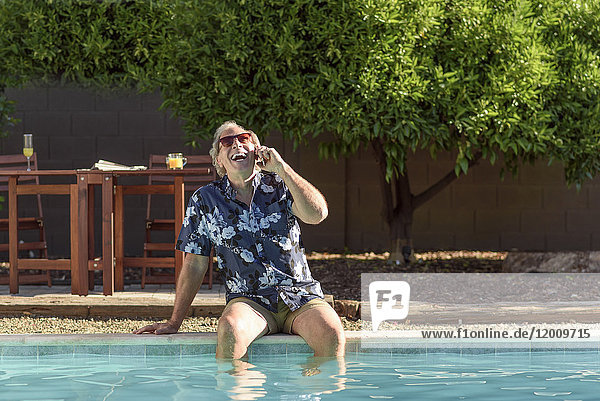 Caucasian man laughing on cell phone with legs in swimming pool