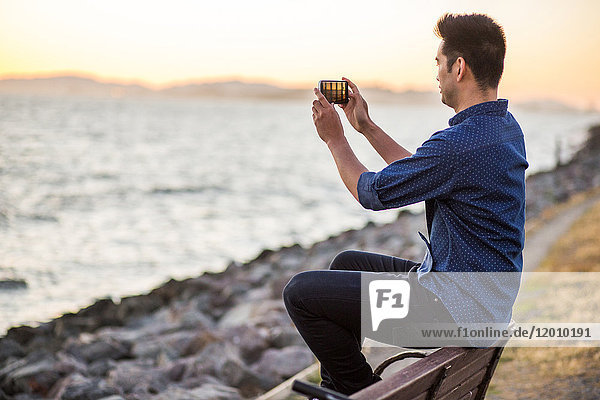 Chinese man sitting on bench photographing ocean with cell phone