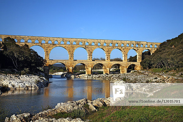 Frankreich  Südfrankreich  Pont du Gard  römisches Aquädukt aus dem I. Jahrhundert am Fluss Gardon. Gesamtansicht mit dem Gardon-Fluss  felsigem Ufer  keine Touristen und blauem Himmel.
