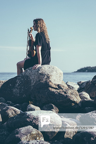 Flachwinkelansicht einer Frau mit Geige  die auf Felsen am See sitzt und dabei wegschaut