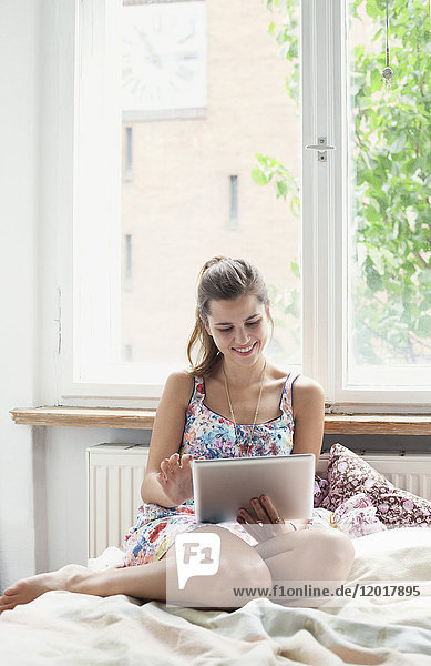 Schöne Frau mit digitalem Tablett  während sie zu Hause auf dem Bett gegen das Fenster sitzt.