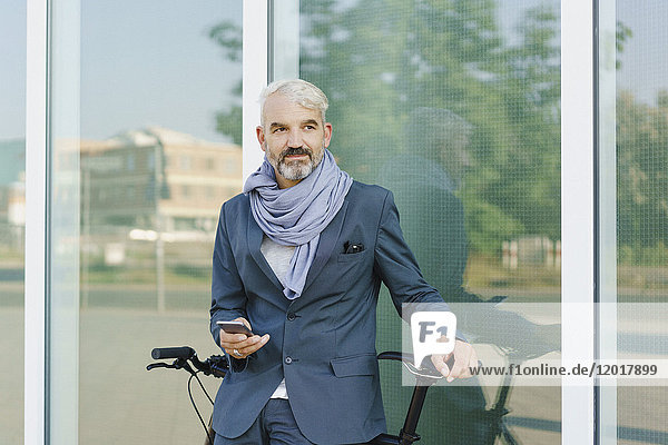 Businessman with bicycle holding smart phone while standing against glass wall