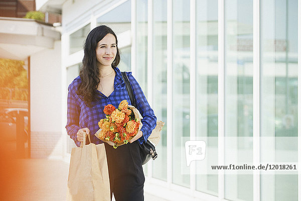 Lächelnde Frau hält Blumenstrauß und Papiertüte  während sie in der Stadt steht.