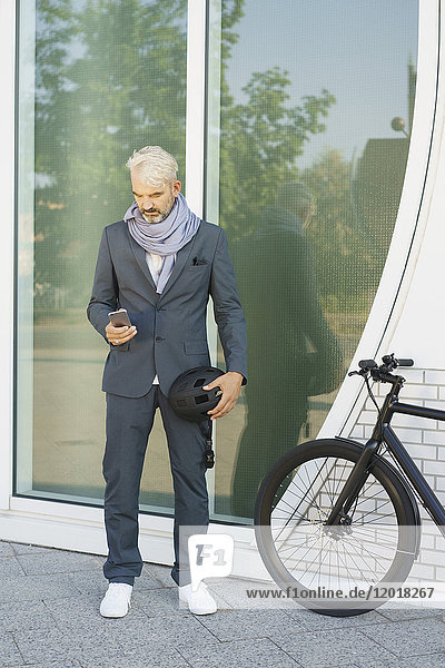 Businessman using mobile phone while standing by bicycle against glass wall