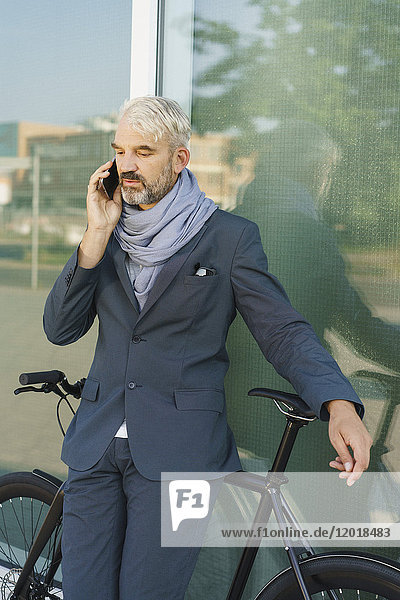 Businessman with bicycle talking on mobile phone while standing against glass building