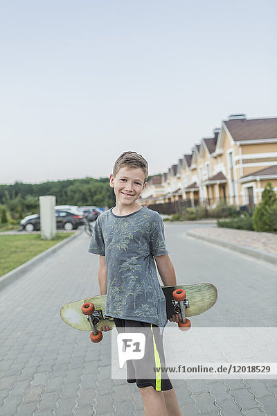 Portrait of smiling boy holding skateboard while standing on street against clear sky