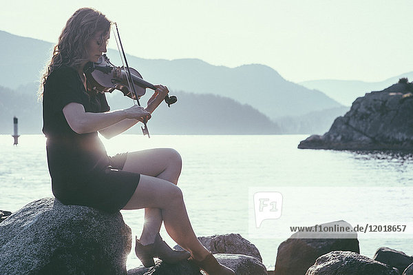 Backlit woman playing violin while sitting on rock at lakeshore against mountains