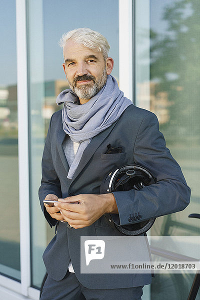 Portrait of confident businessman holding mobile phone and cycle helmet while standing against glass