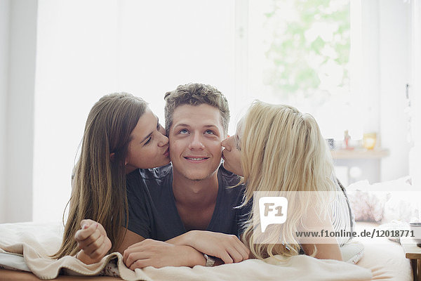 Young female friends kissing handsome man lying on bed at home