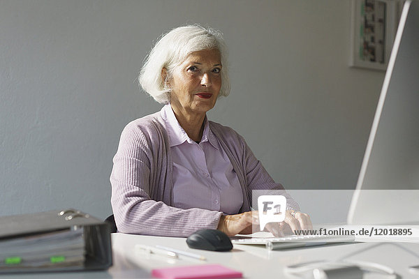 Porträt einer älteren Frau  die im Büro an der Wand sitzt und den Computer benutzt.