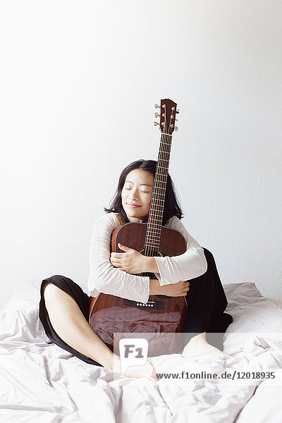 Lächelnde Frau mit geschlossenen Augen  die die Gitarre umarmt  während sie zu Hause auf dem Bett sitzt.